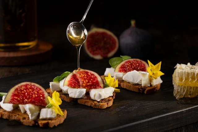 Fig and Goat's Cheese canapés, being drizzled with honey. Set on a wooden board, against a dark background.