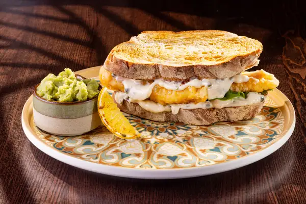 A toasted fish finger sandwich filled with mushy peas and tartare sauce, served with a pot of mushy peas and a charred lemon wedge.