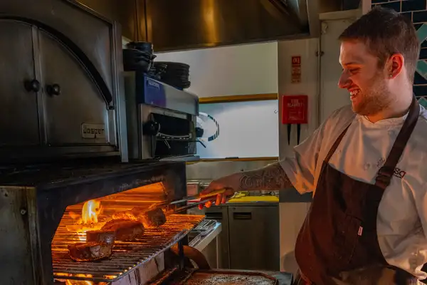 A chef uses metal tongs to turn several steaks over open flames inside a Josper grill in the Cocina kitchen.