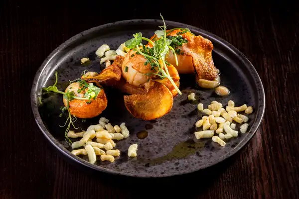Pan-seared scallops are plated with crisp pieces of pork belly, pale artichoke purée, green oil and small leaves.