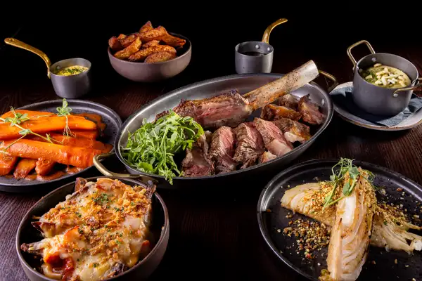 A sliced tomahawk steak rests in an oval pan with rocket, surrounded by bowls of carrots, potato, broccoli cheese, cabbage and sauces.