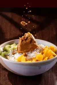 A white bowl of açaí topped with mango cubes, kiwi slices, granola, coconut flakes and Biscoff sprinkle on a dark table.