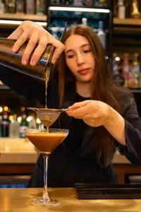 A bartender double-strains a dark espresso martini into a coupe, forming a pale layer of foam on the surface.