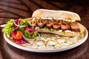 A toasted ciabatta sandwich filled with sliced pink steak, caramelised onions, garlic aioli and rocket, served with a side salad.