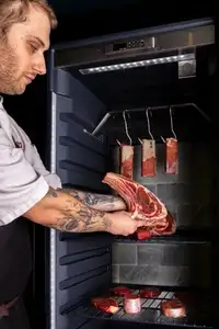 A chef places a large raw bone-in beef cut inside a lit dry-ageing cabinet containing hanging and shelved pieces of beef.