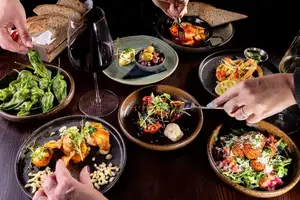 Diners reach across a table filled with tapas dishes, bread and wine glasses, including prawns, padrón peppers, calamari, cauliflower and scallops.