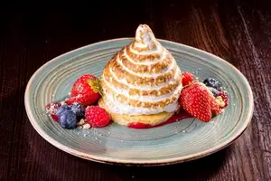 A tall spiral of torched meringue stands on a round dessert base with strawberries, raspberries, blueberries, crumbs and red sauce.