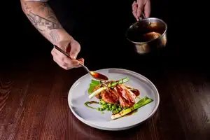 A chef pours sauce over a sliced rack of lamb served with a lamb croquette, peas, broad beans and charred baby leek.