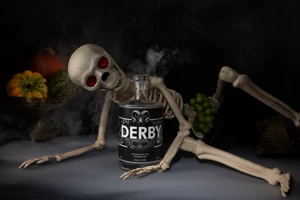 A bottle of Withers Derby Gin lies beside a reclining skeleton surrounded by candles, cobwebs and autumn leaves in a dark Halloween scene.
