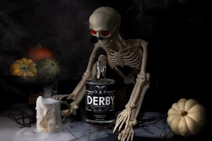 A bottle of Withers Derby Gin stands beside a skeleton holding a candle, surrounded by cobwebs, autumn leaves and Halloween decorations.