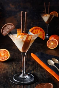 The foreground dessert also has orange peel and a chocolate-orange segment, with a second coupe, halved oranges, a dark napkin and spoons behind it.