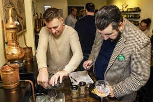 One participant pours ingredients into a small glass while the other uses a spoon beside jars and a bowl, with other guests visible behind them.
