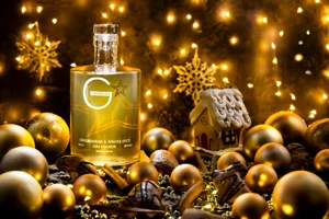 A bottle of Withers Gingerbread & Winter Spice Gin Liqueur stands beside a decorated gingerbread house with Christmas lights and festive decorations.
