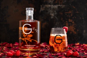 Fresh raspberries and scattered rose petals cover the dark reflective surface around the bottle and ice-filled glass.