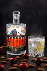 The bottle and ice-filled glass sit on a dark reflective surface beneath a smoky black background, with the red fruit and pale nuts across the foreground.