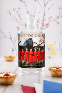 A bottle of Withers Victorious Cherry Bakewell Gin stands beside two Bakewell tarts with cherries and scattered almonds.