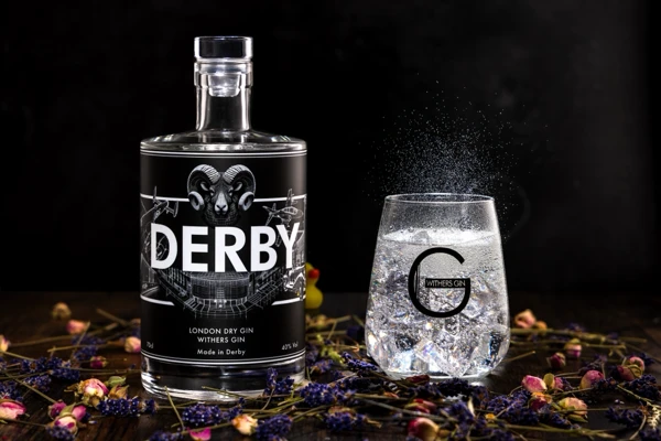 A bottle of Withers Derby Gin stands beside a gin and tonic in a branded glass, surrounded by juniper berries and fresh botanicals.