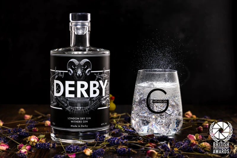A bottle of Withers Derby Gin stands beside a gin and tonic in a branded glass, surrounded by juniper berries and fresh botanicals.