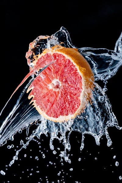 Pink grapefruit half surrounded by curling sheets of water and suspended droplets against a black background.