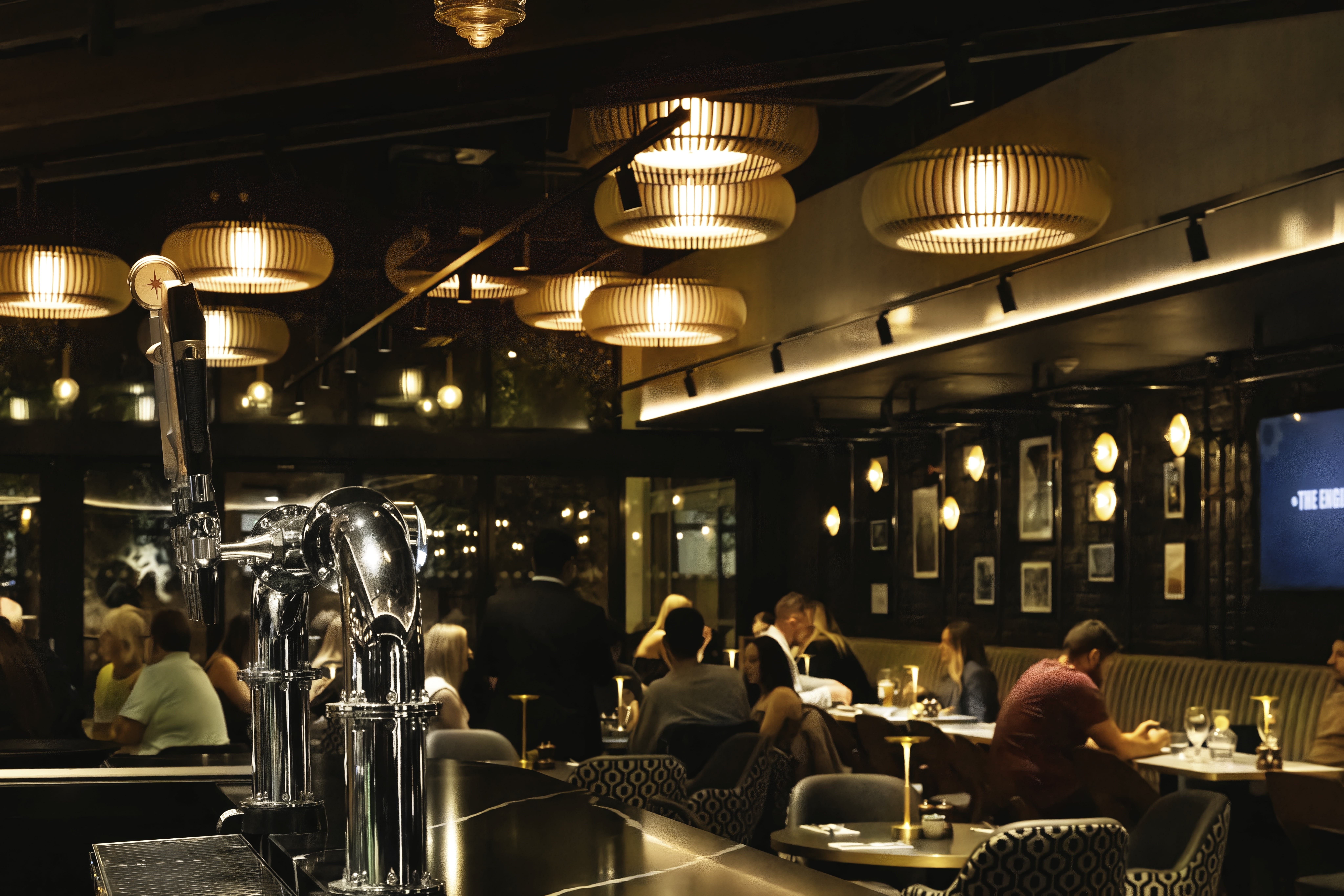 Chrome beer taps stand in the foreground of The Engine Room, with occupied dining tables and pendant lights beyond.
