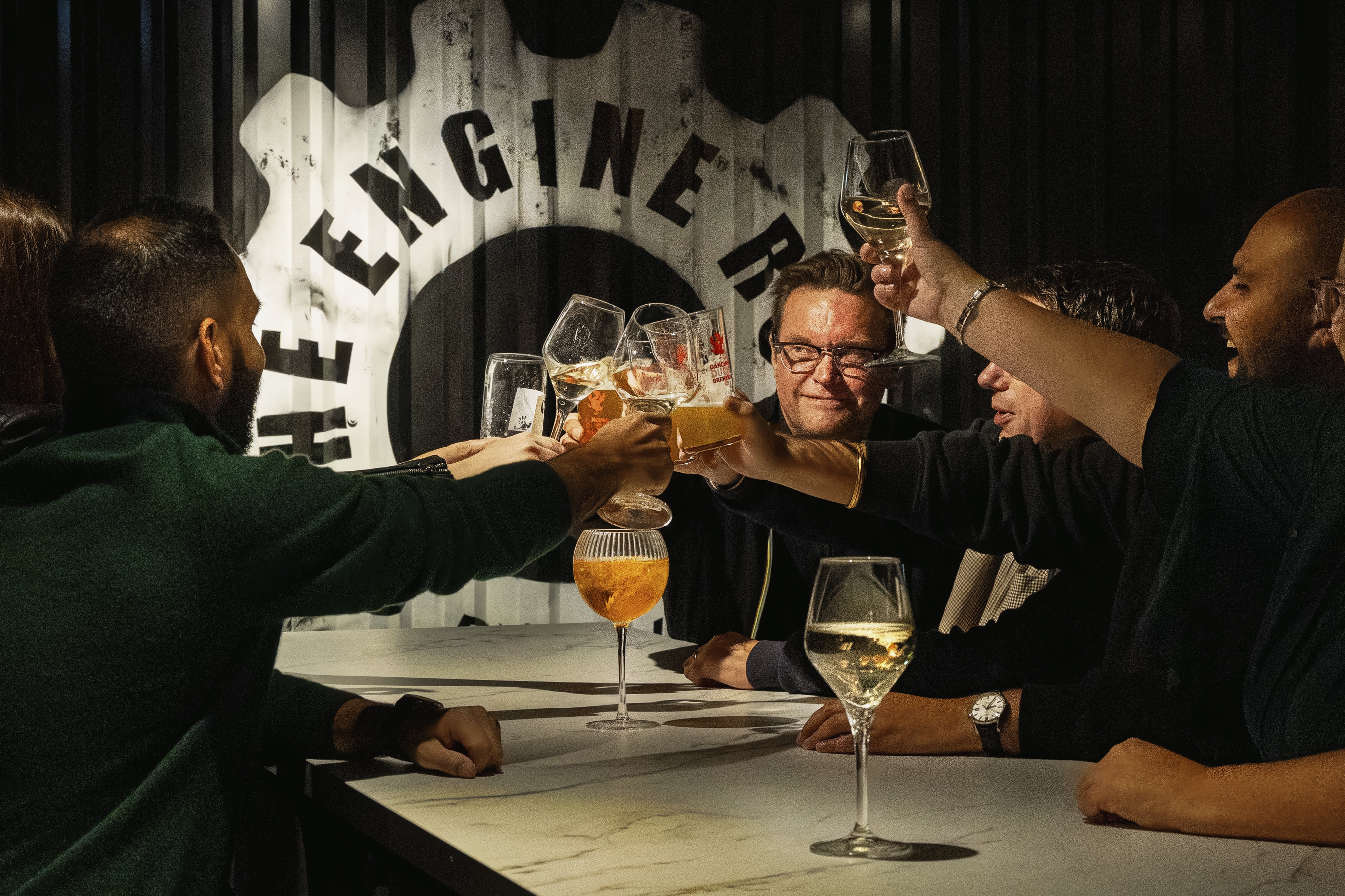 Five diners raise beer, wine and mixed drinks around a white table beneath The Engine Room gear logo.