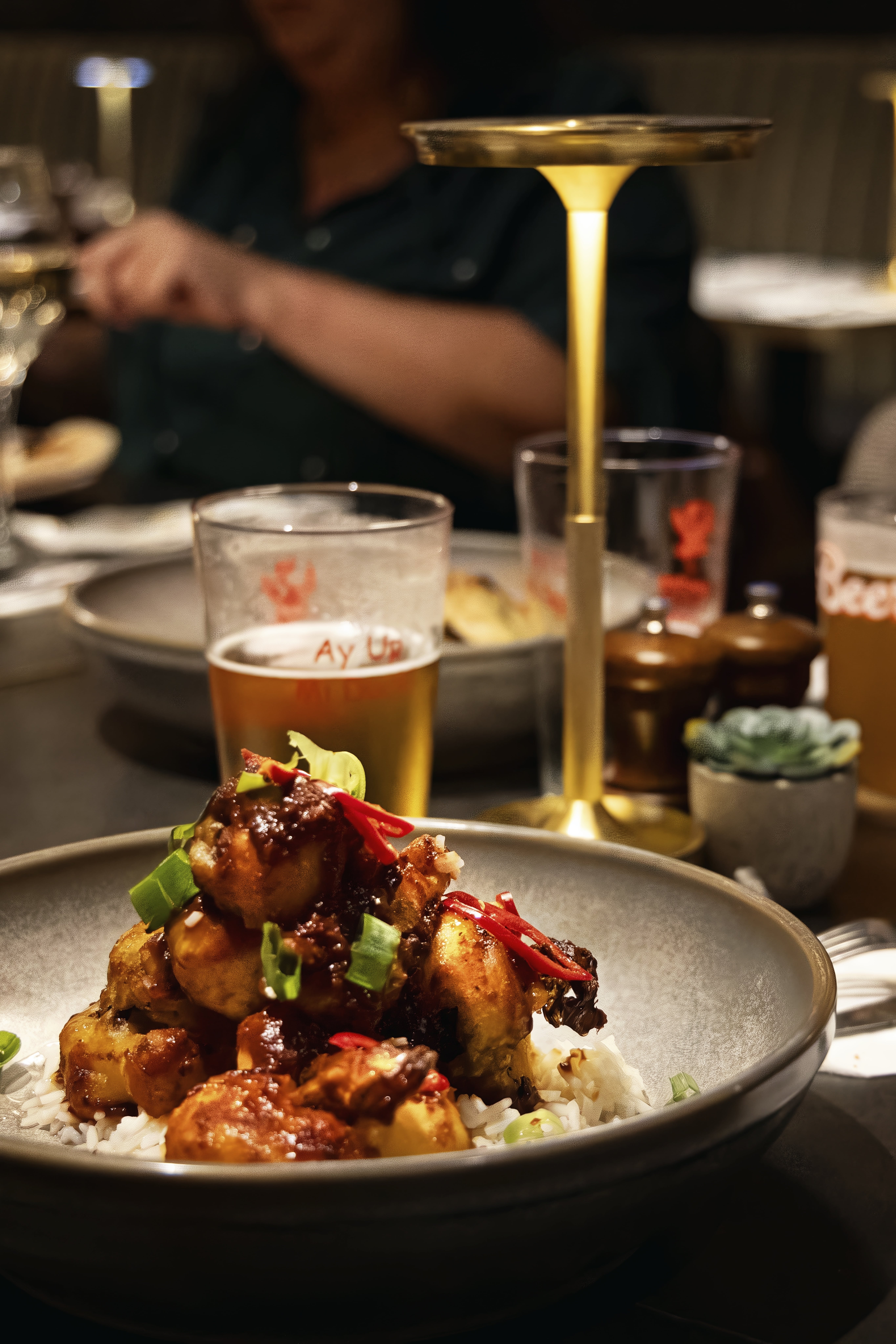 A glazed dish is served over white rice and finished with sliced red chilli and spring onion, with beer and a glowing table lamp behind it.