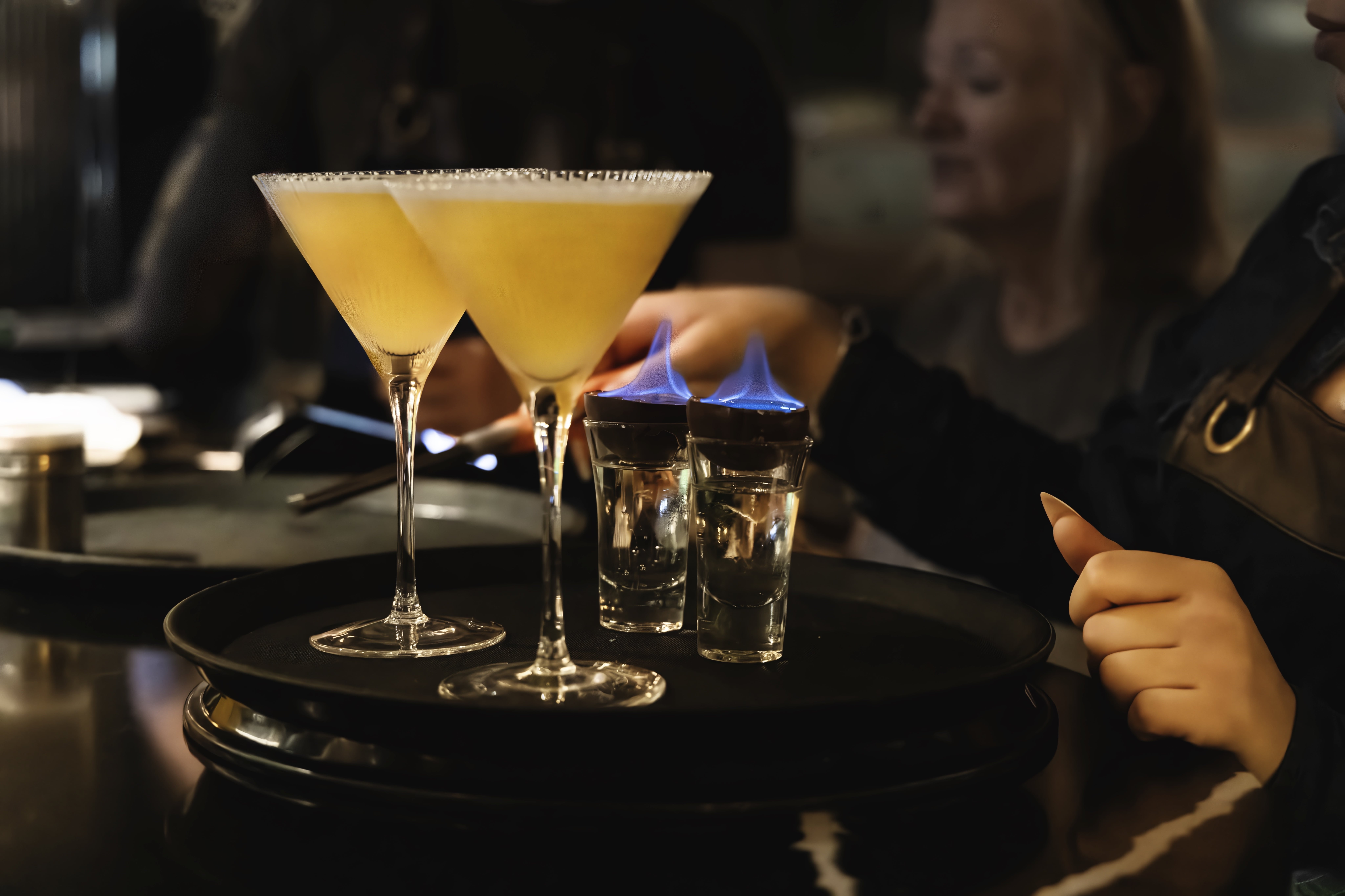 Two Passionfruit Martinis and two Flaming Sambuca shots sit on stacked black trays beside a diner.