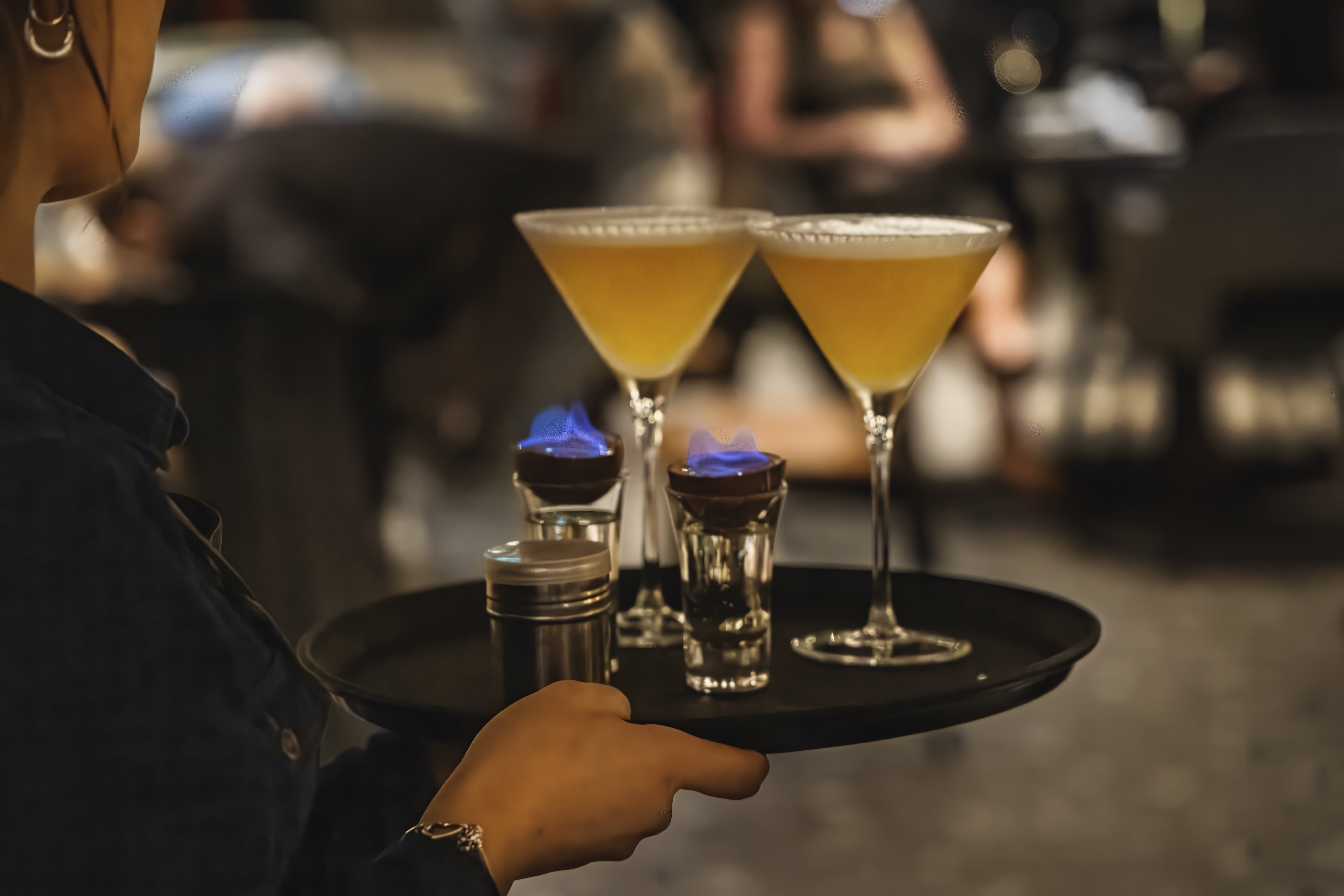 A server carries two Passionfruit Martinis and two Flaming Sambuca shots through the restaurant on a black tray.