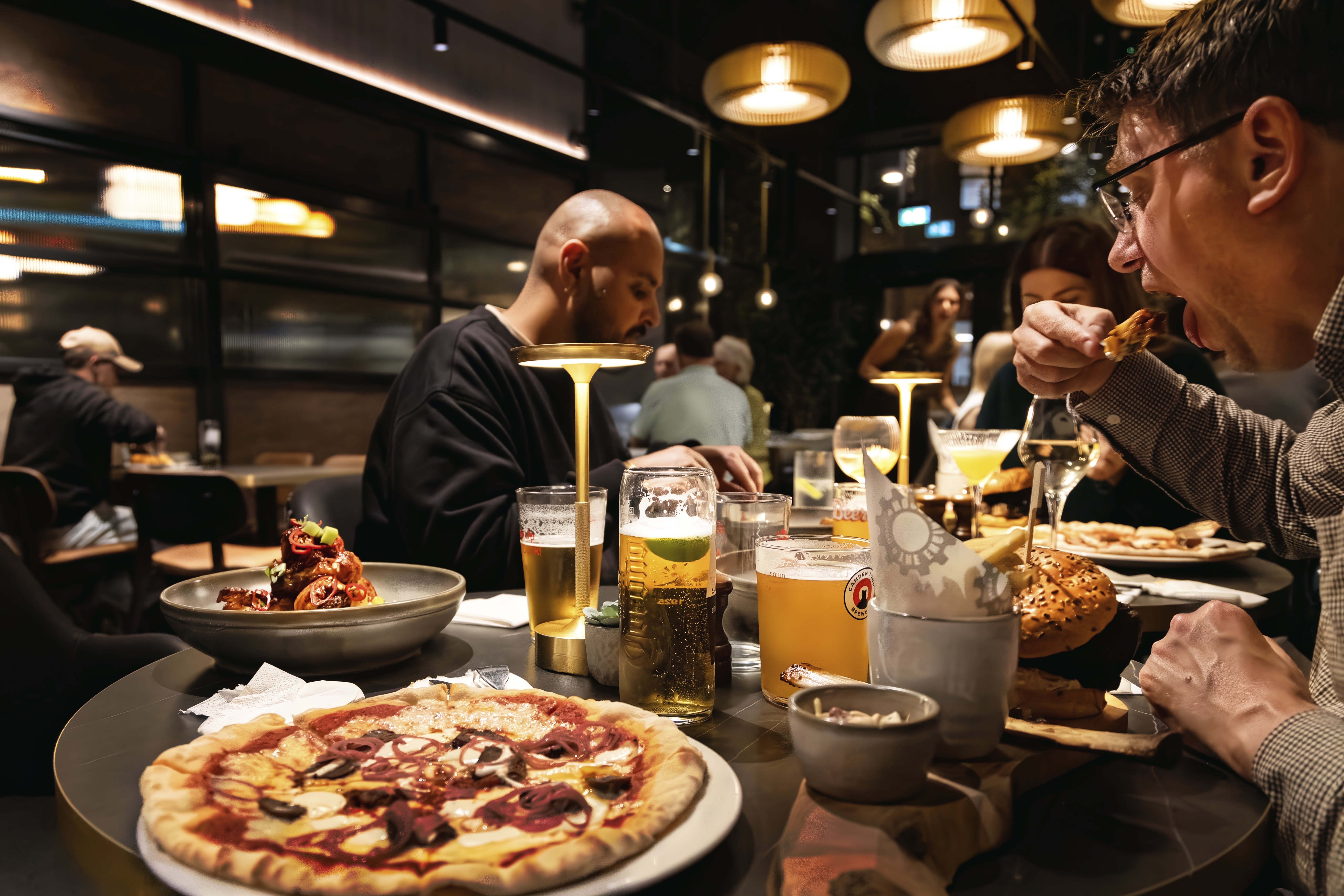 Diners eat around a table holding a pizza, a bone-in burger, another plated dish and several beers.