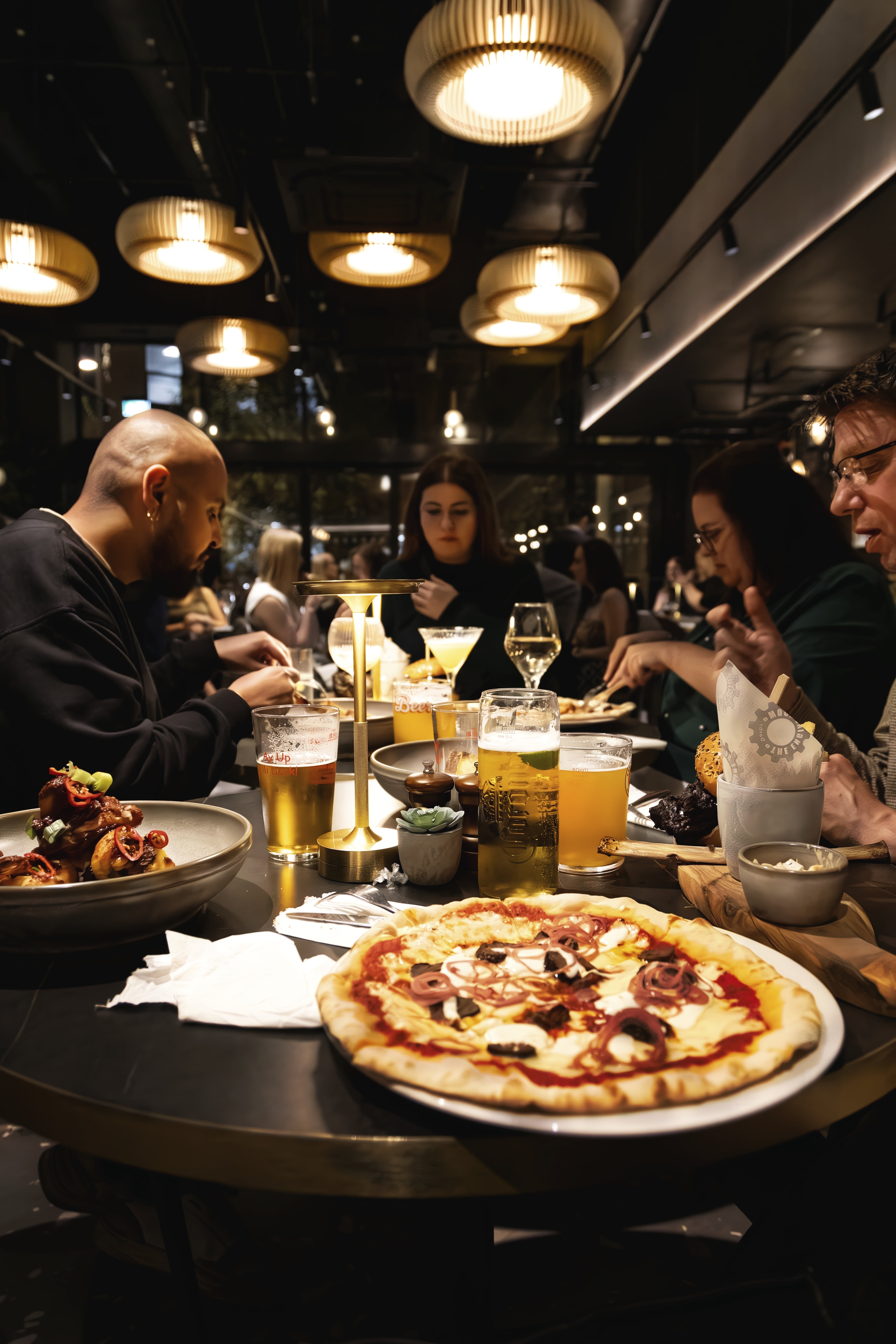 A pizza fills the foreground as a group eats around a crowded table with beer, wine and other dishes.