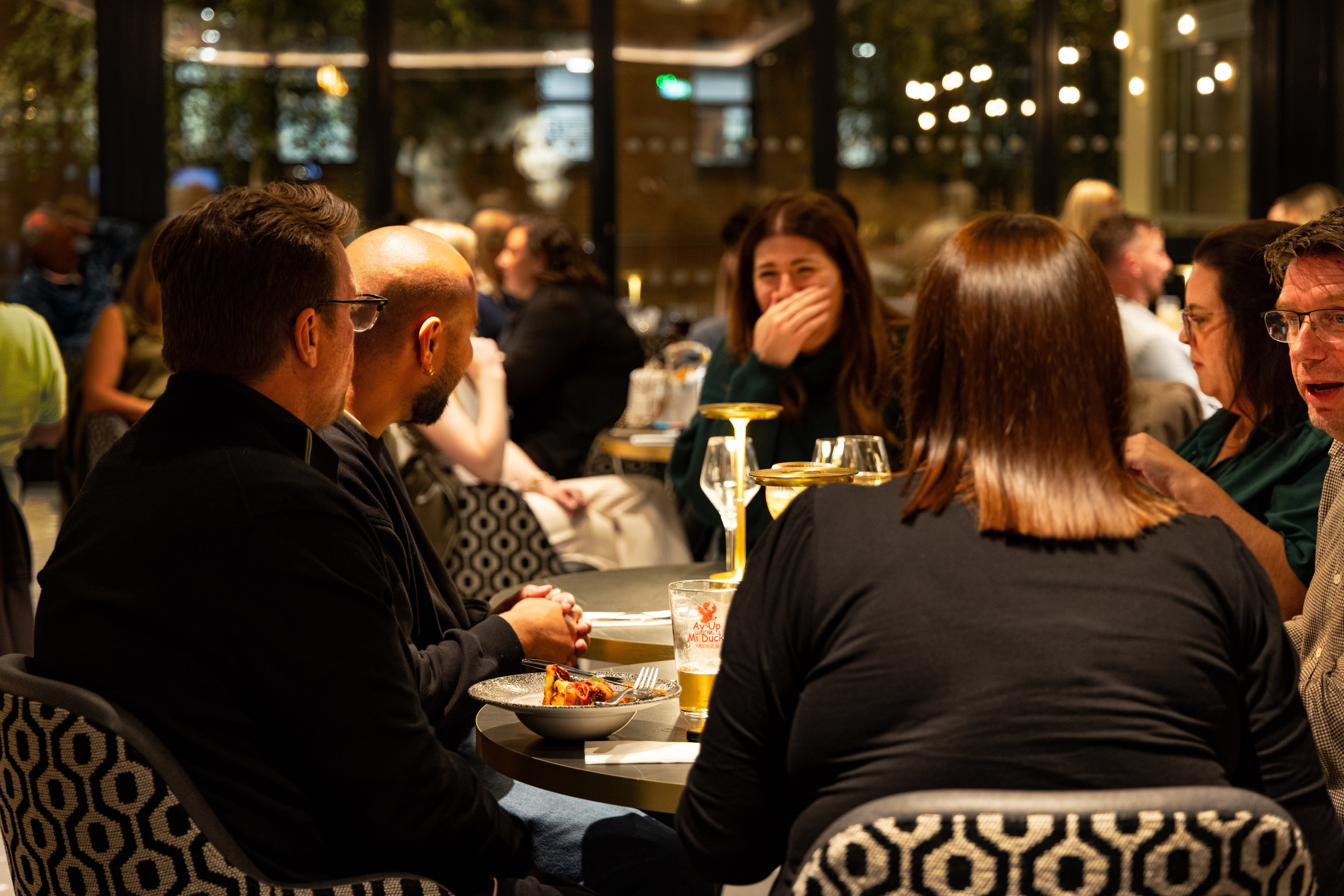 Several diners talk around a table set with plates, beer glasses and water in a busy restaurant.