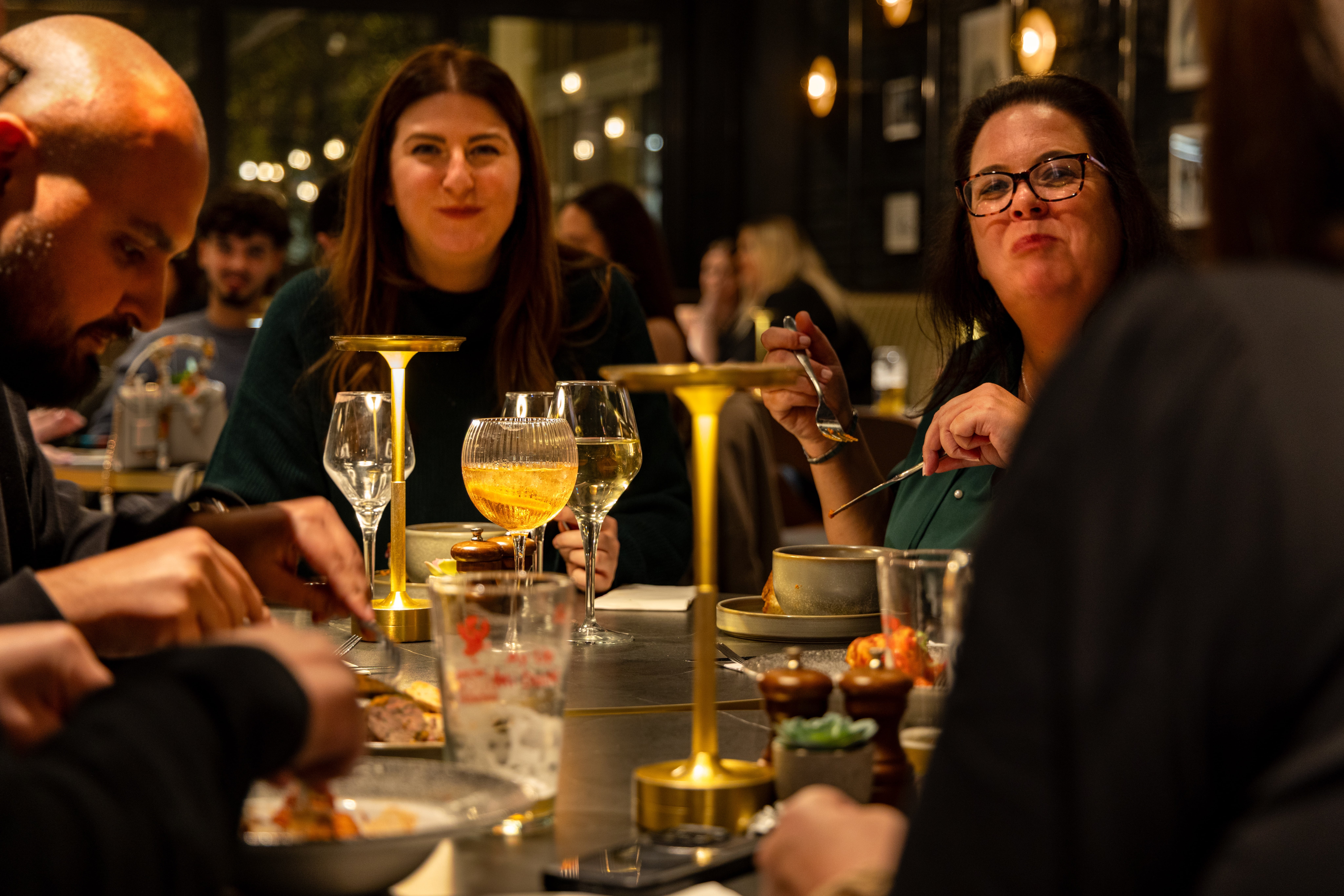 Two women face the camera across a dining table lit by a small brass lamp, with plates and drinks around them.