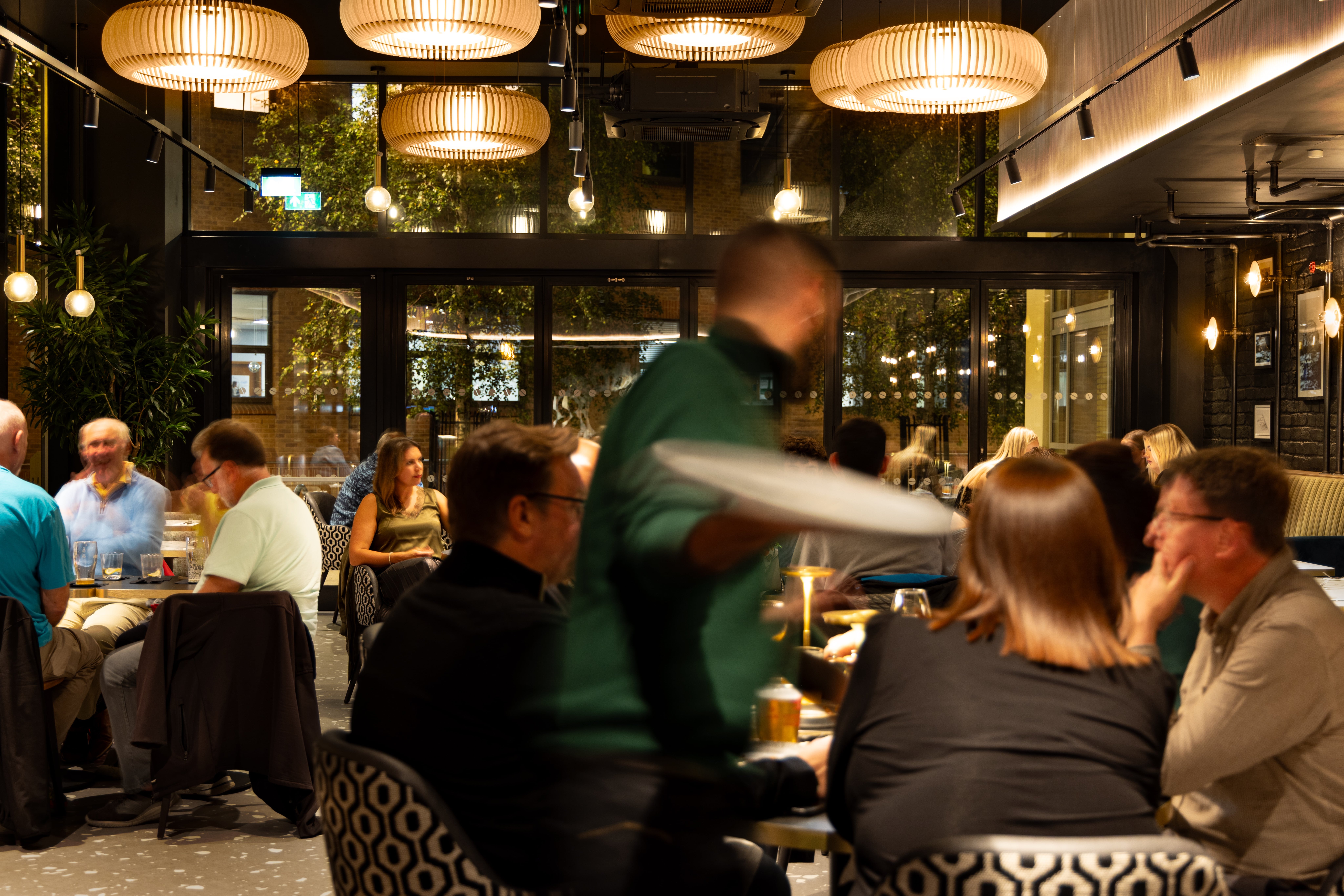 A waiter moves between closely spaced restaurant tables while carrying plates to diners.