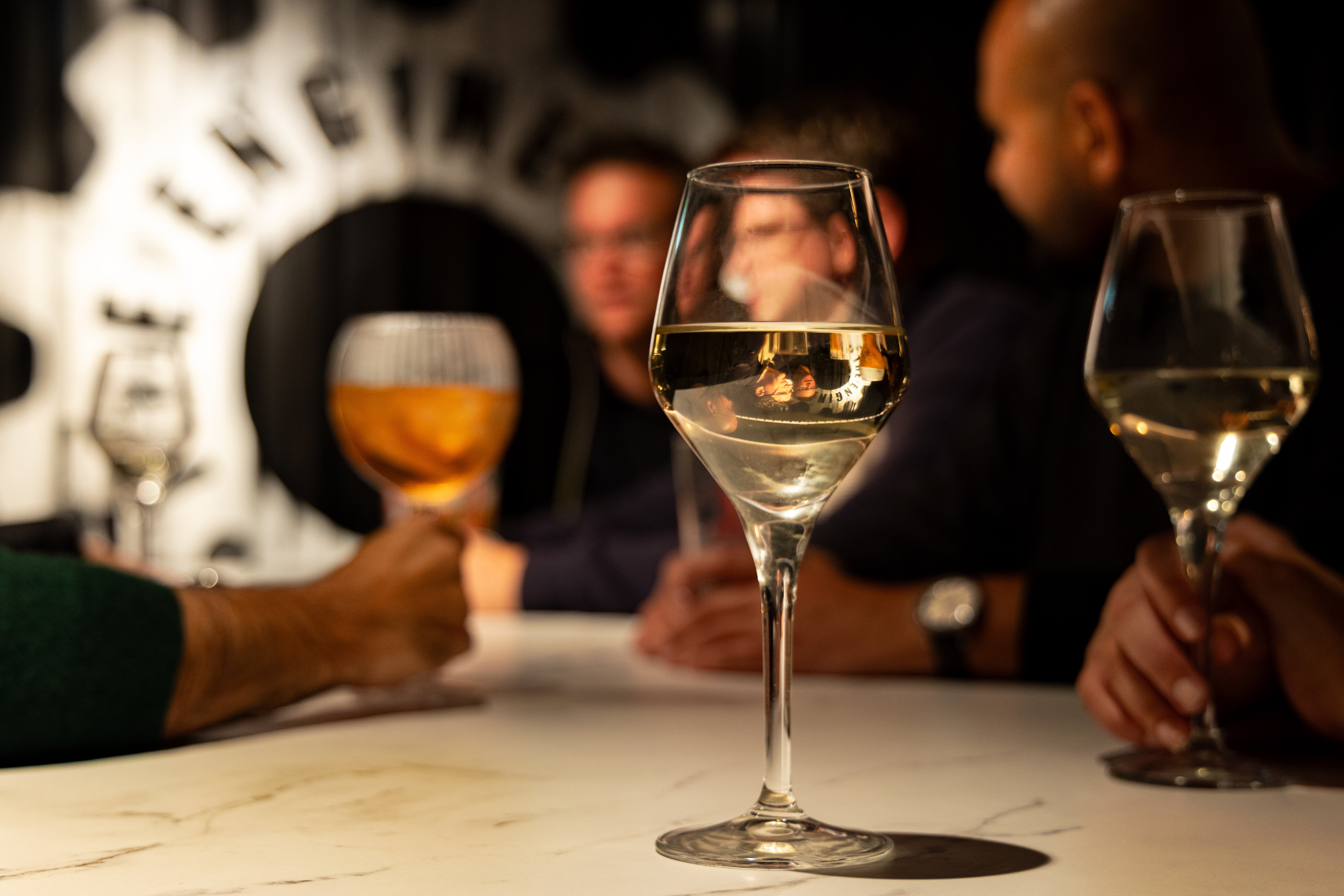 A glass of white wine stands in sharp focus on a restaurant table, with diners and other drinks blurred behind it.