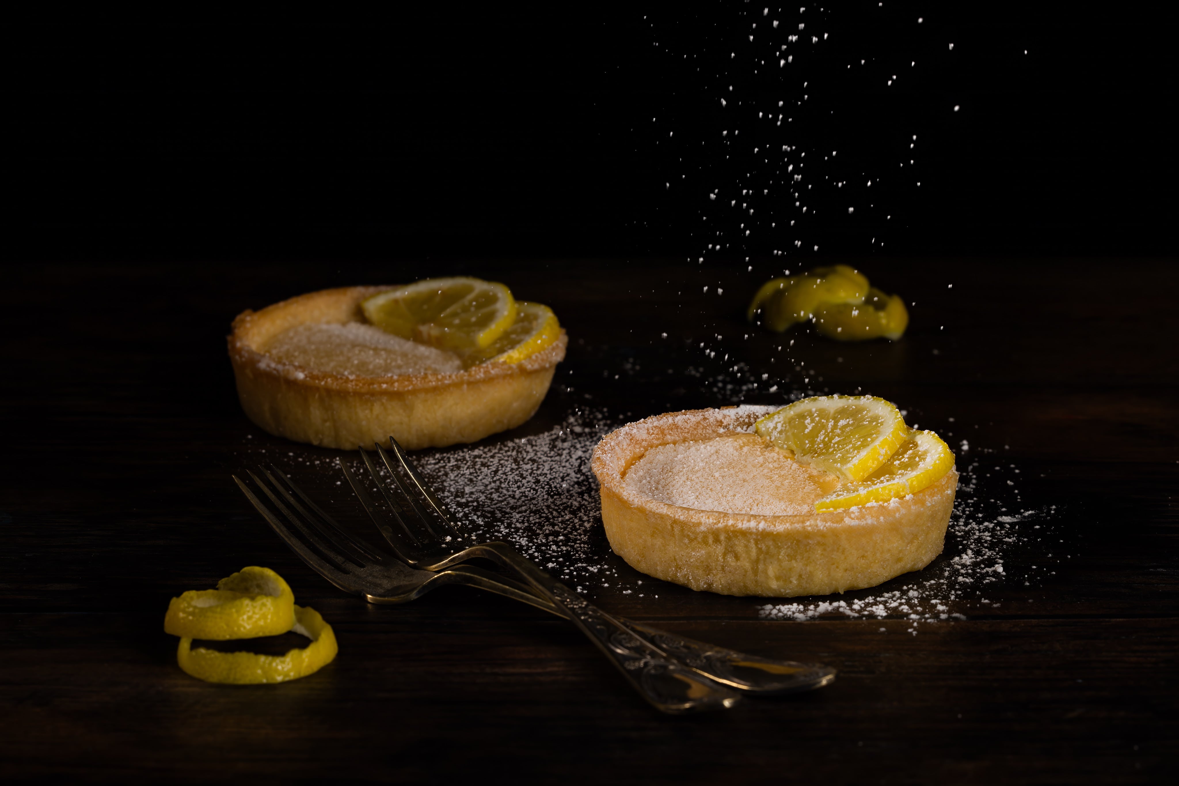 Creative photograph of two lemon tarts, with sprinkling icing sugar and lemon peel. Dramatic Food Photograph by JaxMore Food and Drink Photography, shot in their Derby studio.